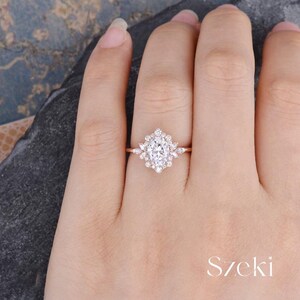 Pu&ograve; includere: Un anello di fidanzamento in oro rosa con una pietra centrale ovale circondata da diamanti pi&ugrave; piccoli. L'anello ha un delicato design floreale.