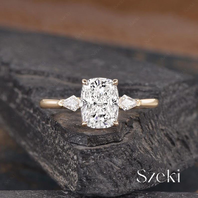 K&ouml;nnte beinhalten: Ein goldener Ring mit einem gro&szlig;en Diamanten im Kissenschliff als Mittelst&uuml;ck, flankiert von zwei kleineren, dreieckigen Diamanten. Der Ring ist vor einem dunklen, strukturierten Hintergrund platziert. Das Wort "Szeki" ist unten rechts sichtbar.