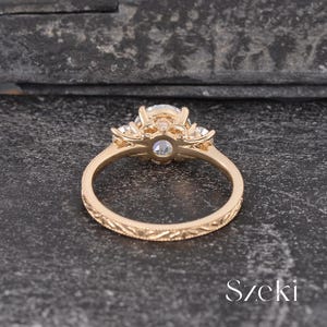 May include: A gold engagement ring with a large, clear center stone and smaller side stones. The ring has a detailed, engraved band and a decorative setting. The ring is set against a dark, textured background.