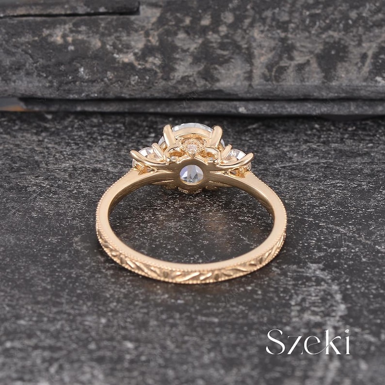 May include: A gold engagement ring with a large, clear center stone and smaller side stones. The ring has a detailed, engraved band and a decorative setting. The ring is set against a dark, textured background.