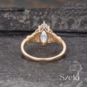 May include: A gold-colored ring with a marquise-cut, clear gemstone. The ring features intricate filigree detailing on the shoulders and around the gemstone setting. The ring is displayed on a dark, textured surface.