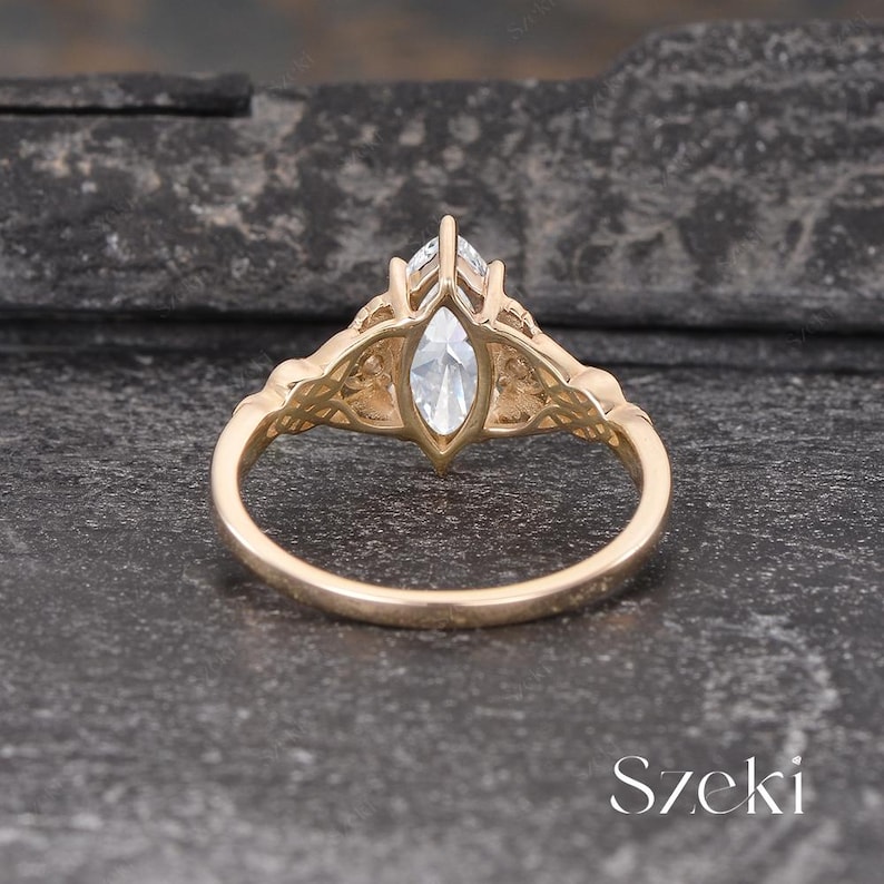 May include: A gold-colored ring with a marquise-cut, clear gemstone. The ring features intricate filigree detailing on the shoulders and around the gemstone setting. The ring is displayed on a dark, textured surface.