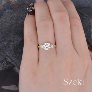 May include: A gold ring with a large, round, clear center stone and two smaller stones on either side. The ring is on a hand, with the word "Szeki" in white script in the lower right corner.