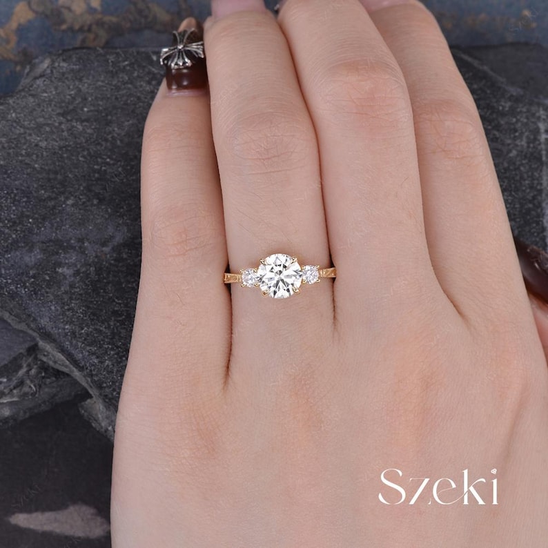 May include: A gold ring with a large, round, clear center stone and two smaller stones on either side. The ring is on a hand, with the word "Szeki" in white script in the lower right corner.