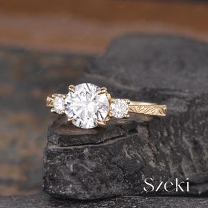May include: A gold engagement ring with a large, round, clear center stone and two smaller side stones. The band features an engraved floral design. The ring is displayed on a dark, textured surface.