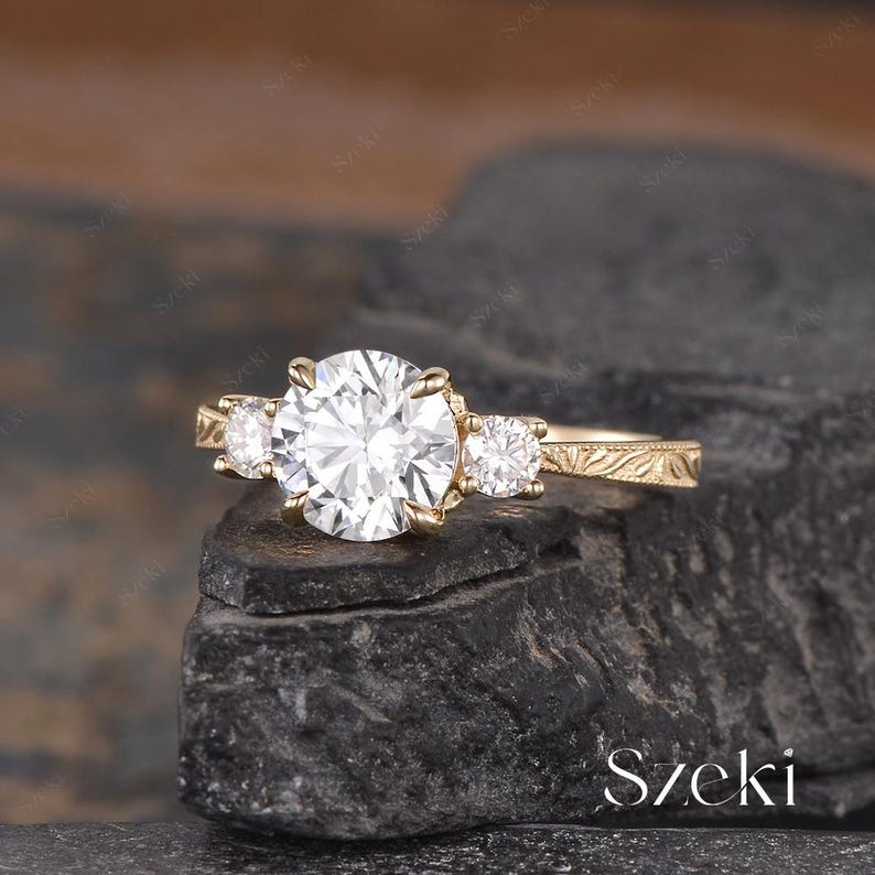 May include: A gold engagement ring with a large, round, clear center stone and two smaller side stones. The band features an engraved floral design. The ring is displayed on a dark, textured surface.