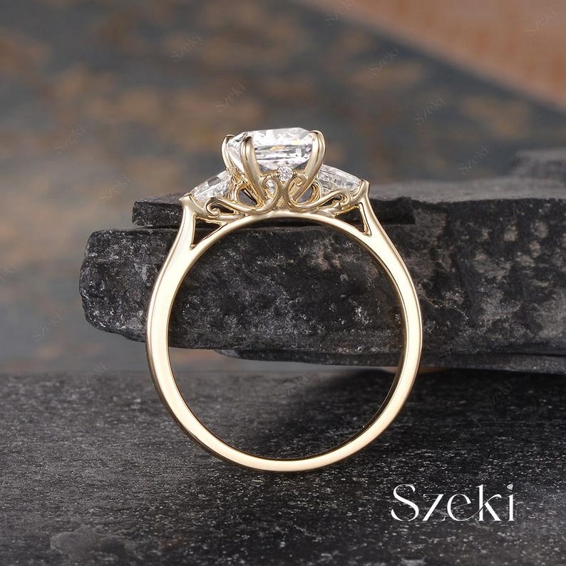 K&ouml;nnte beinhalten: Ein goldener Verlobungsring mit einem gro&szlig;en, rechteckigen Mittelstein und zwei kleineren, trapezf&ouml;rmigen Seitensteinen. Der Ring hat eine filigrane, verzierte Fassung. Das Wort "Szeki" ist sichtbar.