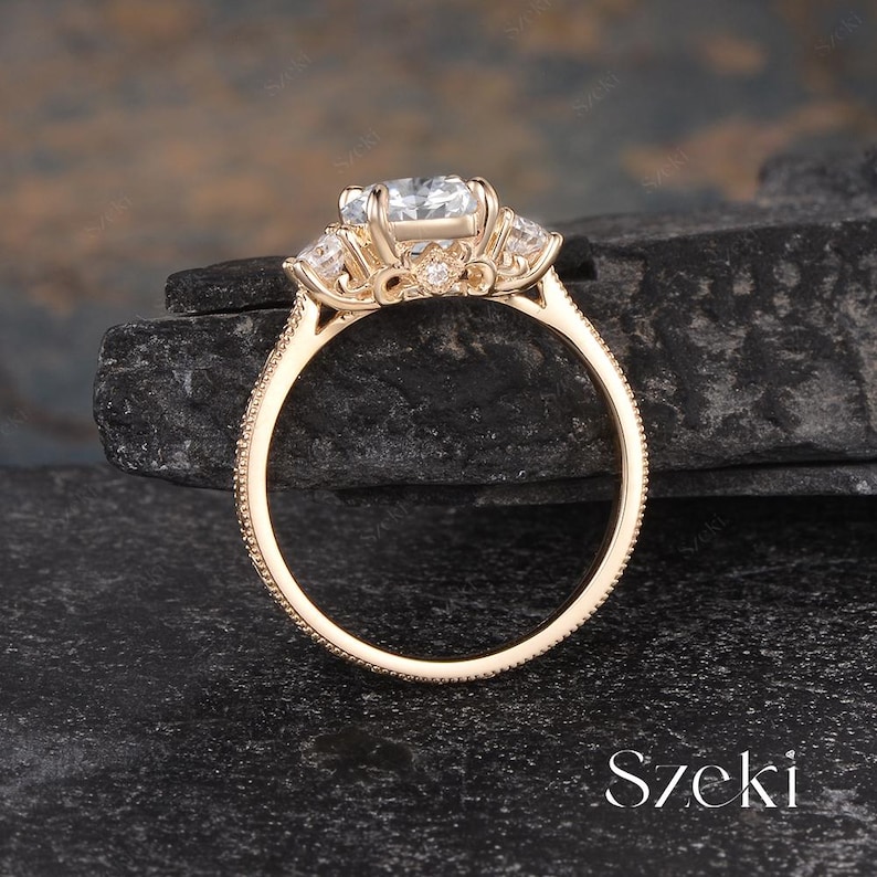 May include: A gold engagement ring with a large, clear center stone and smaller side stones. The ring has a detailed, textured band and is set against a dark, textured background. The word "Szeki" is visible.