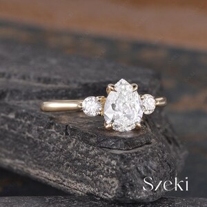 May include: A gold engagement ring featuring a large, pear-shaped diamond as the centerpiece, flanked by two smaller round diamonds. The ring is set on a dark, textured surface. The word "Szeki" is visible in the bottom right corner.
