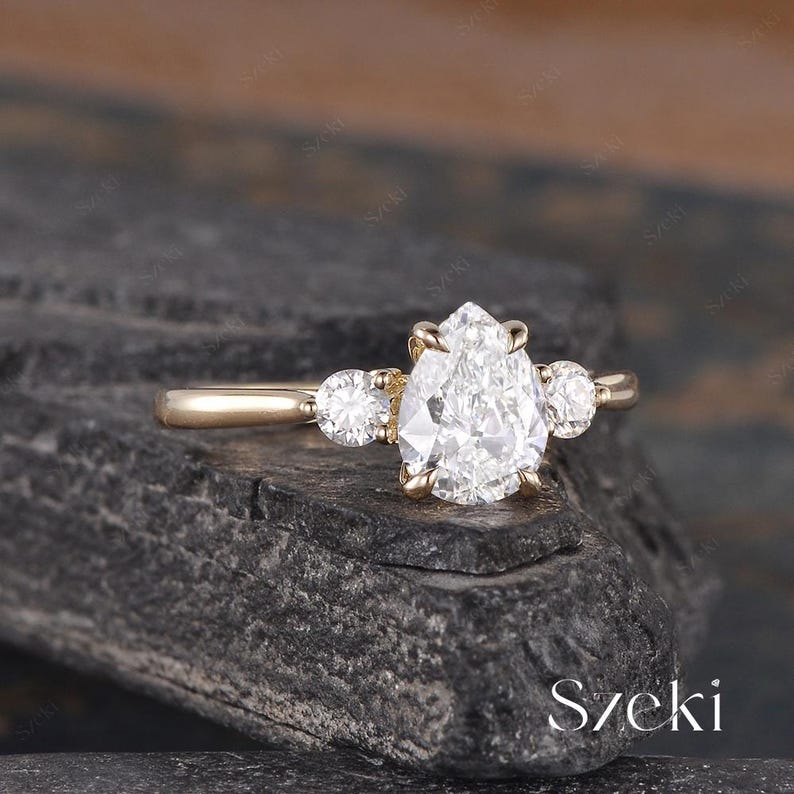 May include: A gold engagement ring featuring a large, pear-shaped diamond as the centerpiece, flanked by two smaller round diamonds. The ring is set on a dark, textured surface. The word "Szeki" is visible in the bottom right corner.