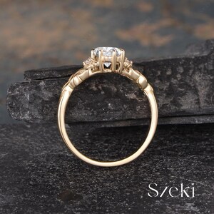 May include: A gold engagement ring with a large, clear gemstone at the center. The ring has a detailed, ornate design with smaller accent stones. The band is a polished gold color. The ring is set against a dark, textured background.