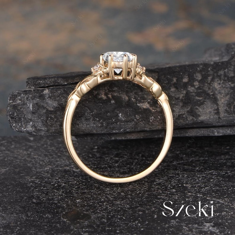 May include: A gold engagement ring with a large, clear gemstone at the center. The ring has a detailed, ornate design with smaller accent stones. The band is a polished gold color. The ring is set against a dark, textured background.