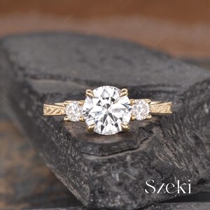 May include: A gold engagement ring with a large, round, clear center stone and two smaller side stones. The band features an engraved design. The ring is set on a dark, textured surface.