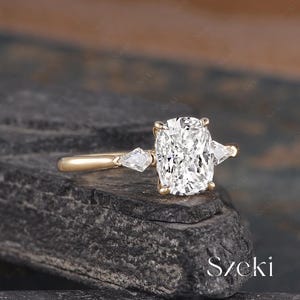 K&ouml;nnte beinhalten: Ein goldener Ring mit einem gro&szlig;en, quadratisch geschliffenen Diamanten in der Mitte und zwei kleineren, birnenf&ouml;rmigen Diamanten auf beiden Seiten. Der Ring liegt auf einer dunklen, strukturierten Oberfl&auml;che. Das Wort "Szecki" ist unten rechts sichtbar.