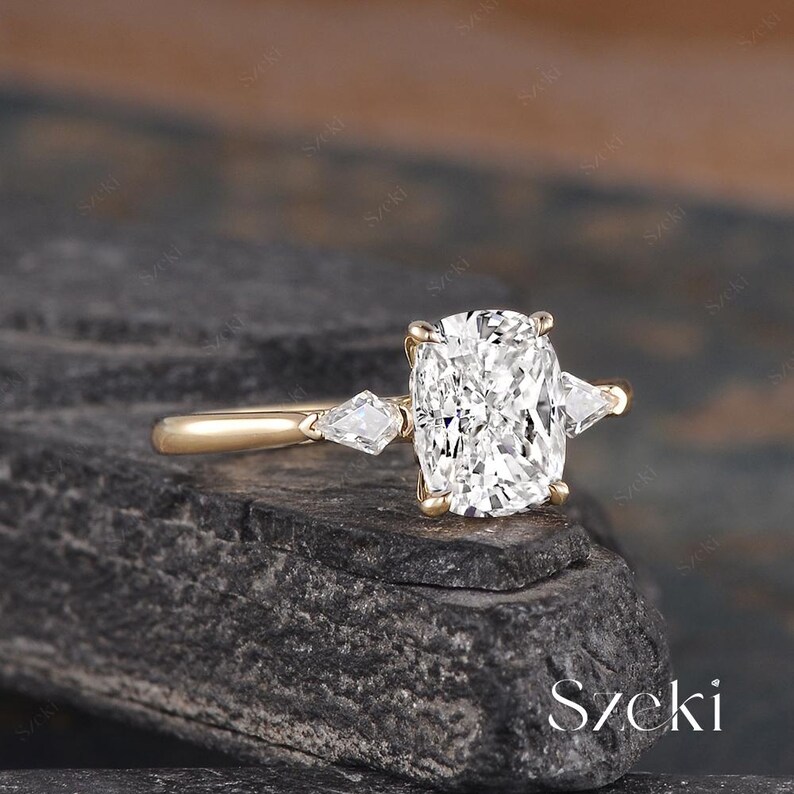 K&ouml;nnte beinhalten: Ein goldener Ring mit einem gro&szlig;en, quadratisch geschliffenen Diamanten in der Mitte und zwei kleineren, birnenf&ouml;rmigen Diamanten auf beiden Seiten. Der Ring liegt auf einer dunklen, strukturierten Oberfl&auml;che. Das Wort "Szecki" ist unten rechts sichtbar.