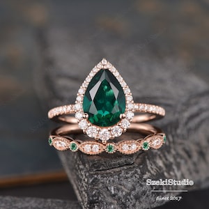 Puede incluir: Un anillo de compromiso de esmeralda en forma de pera engastado en oro rosa con un halo de diamantes más pequeños. La banda de boda a juego presenta un diseño delicado con esmeraldas y diamantes más pequeños.
