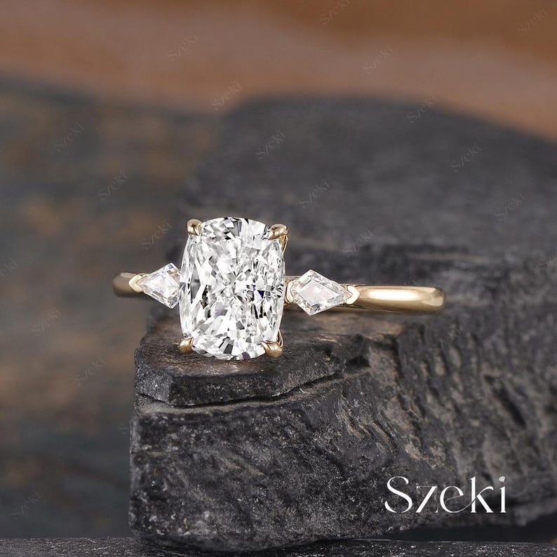 K&ouml;nnte beinhalten: Ein goldener Verlobungsring mit einem gro&szlig;en, rechteckigen, klaren Edelstein als Mittelpunkt. Zwei kleinere, dreieckige Edelsteine flankieren den Hauptstein. Der Ring liegt auf einer dunklen, strukturierten Oberfl&auml;che. Das Wort "Szeki" ist sichtbar.