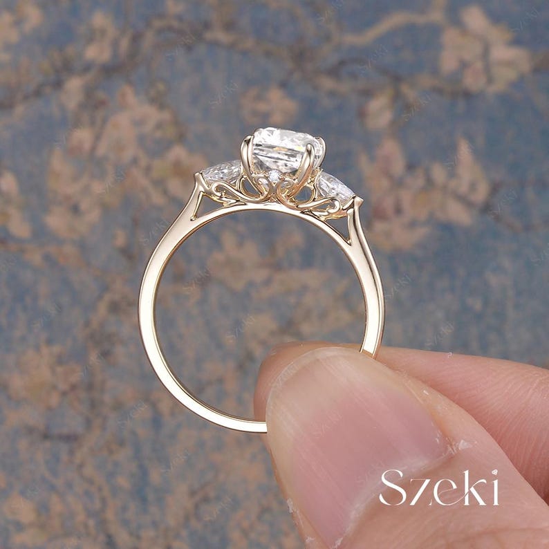 K&ouml;nnte beinhalten: Ein goldener Verlobungsring mit einem gro&szlig;en, quadratisch geschliffenen Mittelstein, flankiert von zwei kleineren Steinen. Der Ring hat ein zartes Band und eine filigrane Fassung. Der Hintergrund ist blau und braun.