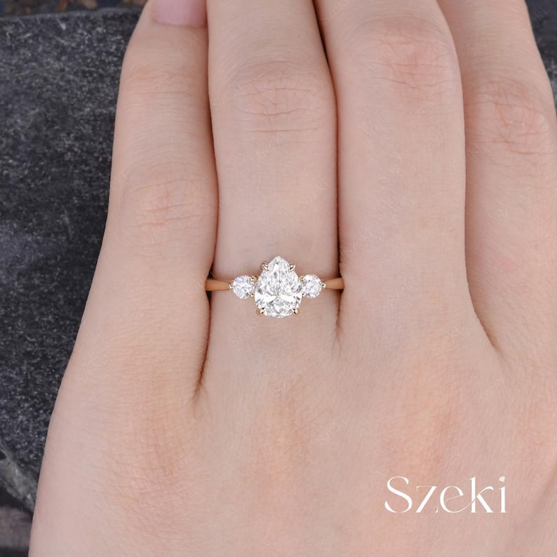 May include: A gold engagement ring with a pear-shaped center stone and two smaller round stones on either side. The ring is set on a delicate band. The word "Szeki" is written in a script font.
