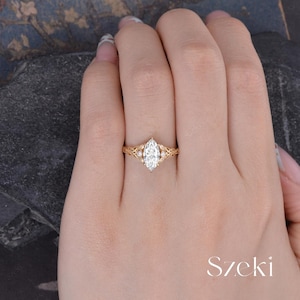 May include: A gold-colored ring with a marquise-cut diamond center stone. The ring features a delicate filigree design with smaller accent stones. The ring is set on a hand.