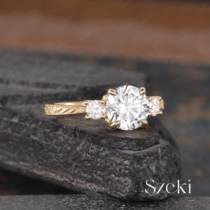 May include: A gold engagement ring with a large, round, clear center stone and two smaller side stones. The ring has an ornate, engraved band. The ring is set on a dark, textured surface.