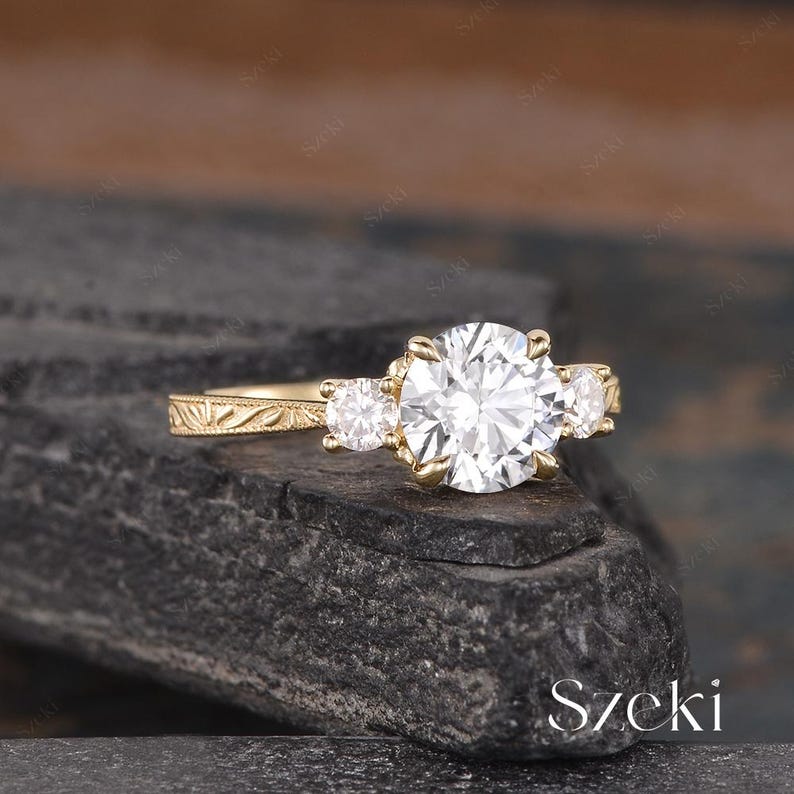 May include: A gold engagement ring with a large, round, clear center stone and two smaller side stones. The ring has an ornate, engraved band. The ring is set on a dark, textured surface.