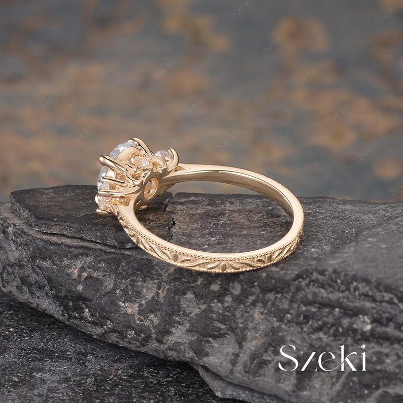 May include: A gold-colored ring with a large, clear gemstone and smaller accent stones. The band features an intricate floral pattern. The ring is set on a dark, textured surface. The word "Szeki" is visible in the image.