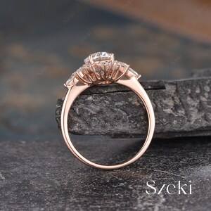 Pu&ograve; includere: Un anello di fidanzamento in oro rosa con una grande pietra centrale rotonda circondata da pietre rotonde pi&ugrave; piccole. L'anello ha una montatura a halo e una fascia con tre pietre a forma di pera su ogni lato.