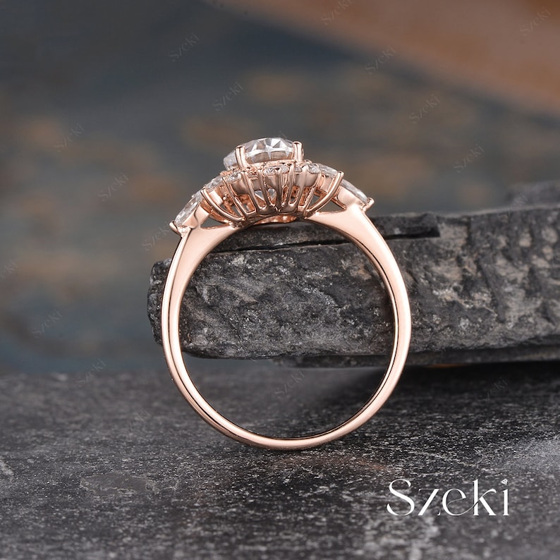 Pu&ograve; includere: Un anello di fidanzamento in oro rosa con una grande pietra centrale rotonda circondata da pietre rotonde pi&ugrave; piccole. L'anello ha una montatura a halo e una fascia con tre pietre a forma di pera su ogni lato.