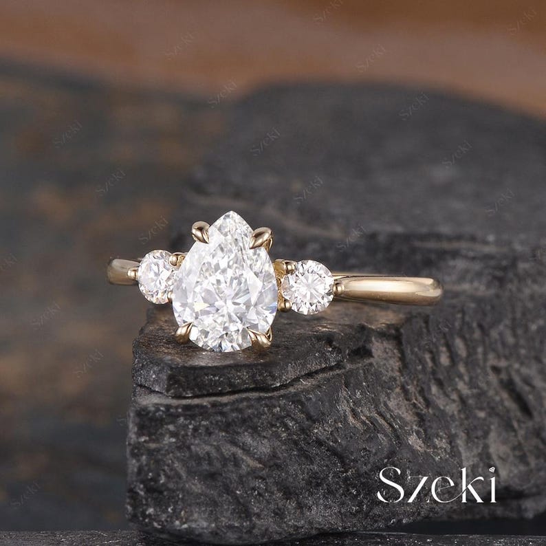 May include: A gold engagement ring featuring a pear-shaped diamond center stone, flanked by two smaller round diamonds. The ring is set on a gold band and is displayed on a dark, textured surface. The word "Szeki" is visible.