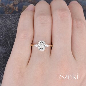 K&ouml;nnte beinhalten: Ein goldener Ring mit einem gro&szlig;en, rechteckigen, klaren Edelstein als Mittelst&uuml;ck, flankiert von zwei kleineren, birnenf&ouml;rmigen Steinen. Der Ring befindet sich an der Hand einer Person. Das Wort "Szeki" ist sichtbar.
