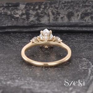 May include: A gold ring with a large, clear, geometric-cut gemstone at the center, flanked by smaller stones. The ring's band is a simple, polished gold. The ring is set against a dark, textured background. The word "Szeki" is visible.