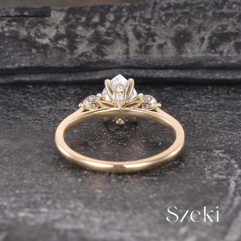 May include: A gold ring with a large, clear, geometric-cut gemstone at the center, flanked by smaller stones. The ring's band is a simple, polished gold. The ring is set against a dark, textured background. The word "Szeki" is visible.