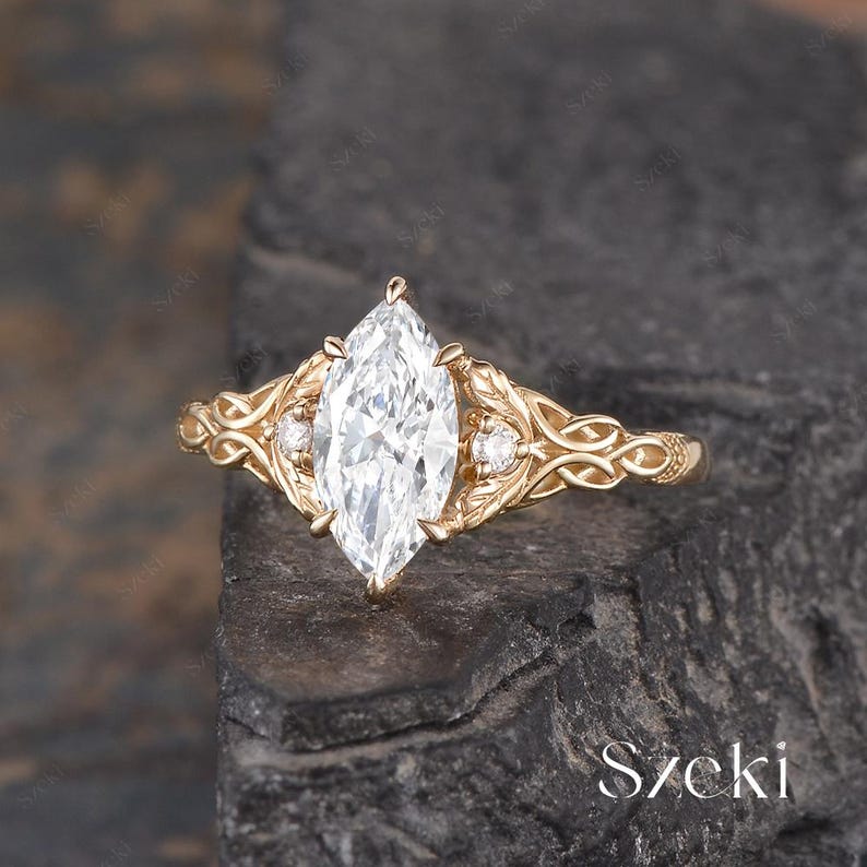 May include: A marquise-cut diamond engagement ring with a gold band. The ring features intricate filigree detailing on the band and around the central diamond, with smaller accent stones on either side. The ring is set against a dark, textured background.