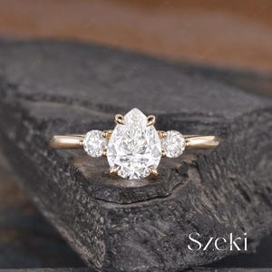 May include: A gold ring featuring a pear-shaped diamond center stone, flanked by two smaller round diamonds. The ring is set on a simple gold band, showcasing a classic and elegant design. The ring is displayed on a dark, textured surface.