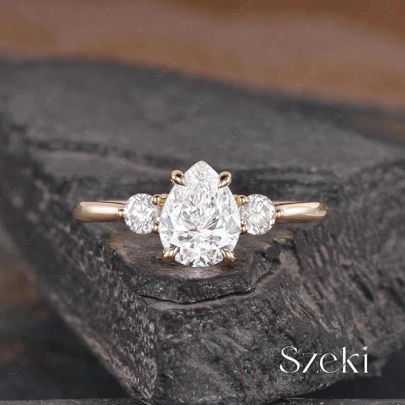 May include: A gold ring featuring a pear-shaped diamond center stone, flanked by two smaller round diamonds. The ring is set on a simple gold band, showcasing a classic and elegant design. The ring is displayed on a dark, textured surface.