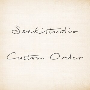 May include: A handwritten sign with the text "Szekistudio Custom Order" in black ink on a white background.
