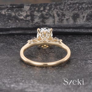 K&ouml;nnte beinhalten: Ein goldener Verlobungsring mit einem gro&szlig;en, klaren Edelstein. Der Ring hat aufwendige Filigranarbeiten um den zentralen Stein und kleinere Steine auf beiden Seiten. Das Band ist glatt und poliert. Das Wort "Szeki" ist sichtbar.