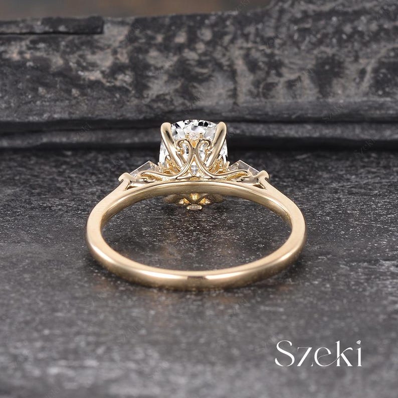 K&ouml;nnte beinhalten: Ein goldener Verlobungsring mit einem gro&szlig;en, klaren Edelstein. Der Ring hat aufwendige Filigranarbeiten um den zentralen Stein und kleinere Steine auf beiden Seiten. Das Band ist glatt und poliert. Das Wort "Szeki" ist sichtbar.