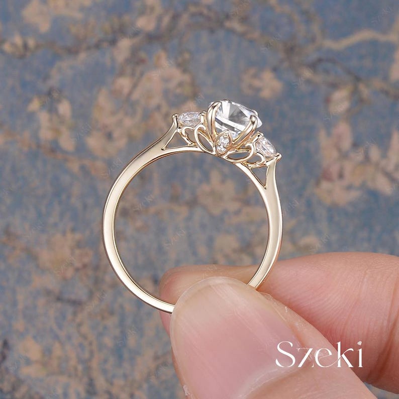 May include: A gold ring with a large, clear, round center stone and two smaller side stones. The ring has a delicate, detailed design around the stones. The ring is held by a person's hand. The word "Szeki" is visible.