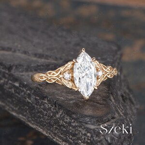 May include: A marquise-cut diamond engagement ring with a gold band. The ring features intricate filigree and leaf details, with smaller accent diamonds on either side of the main stone. The ring is set against a dark, textured background.