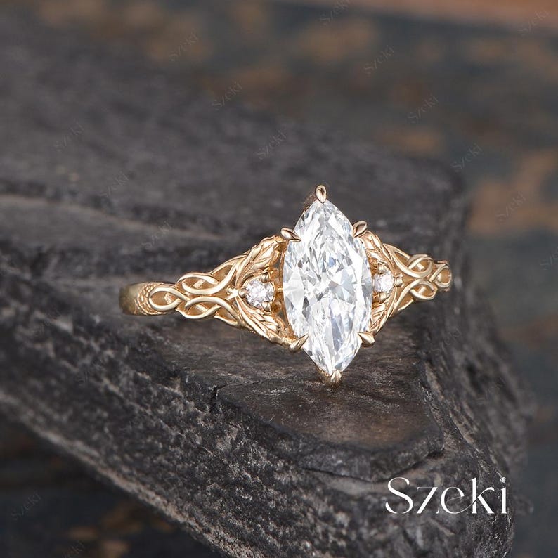 May include: A marquise-cut diamond engagement ring with a gold band. The ring features intricate filigree and leaf details, with smaller accent diamonds on either side of the main stone. The ring is set against a dark, textured background.