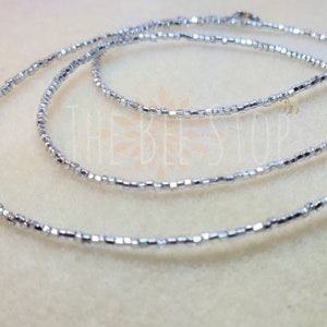 May include: A silver beaded necklace with a clasp closure. The necklace is made of small, square-shaped beads.