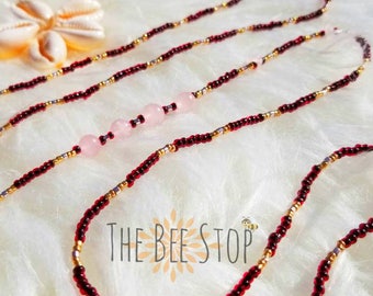 May include: A waist bead necklace with red and gold beads and pink stone accents. The text "THE BEE STOP" is visible in the image.