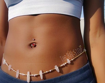 May include: A gold belly chain with white and pink beads and a small red gem. The chain is worn around a person's waist and has the text "THE BEAD STOP" on it.