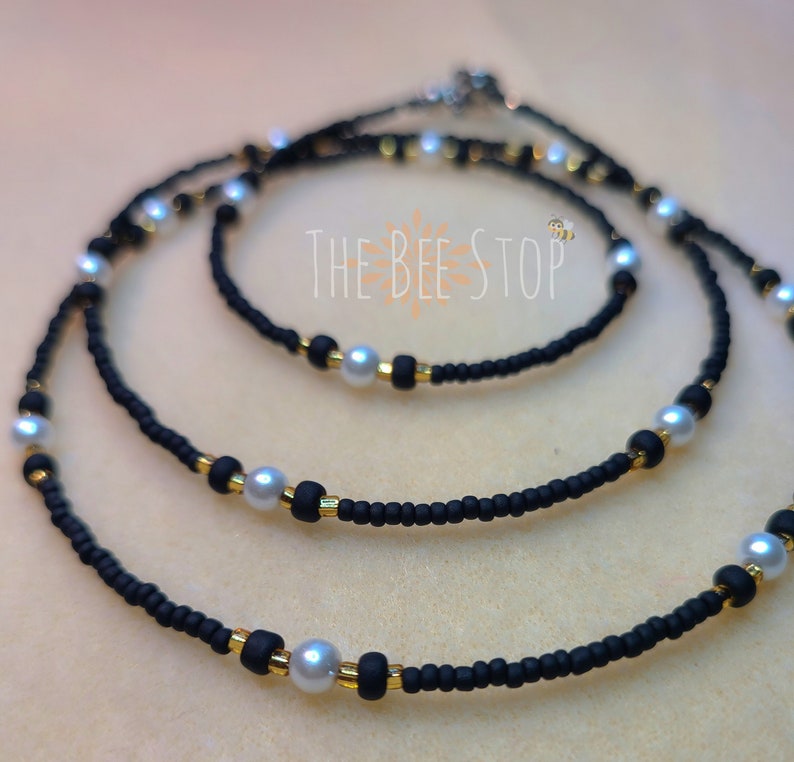 K&ouml;nnte beinhalten: Eine schwarze Perlen-Taillenkette mit goldenen Perlen und wei&szlig;en Perlen. Die Kette ist geschichtet und tr&auml;gt den Text "THE BEE STOP" in der Mitte.
