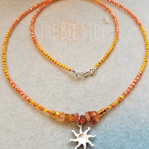 May include: A beaded necklace with orange, yellow, and gold beads. The necklace features a silver sunburst pendant and a cluster of orange and yellow gemstones. The text "THE BEE STOP" is visible in the background.