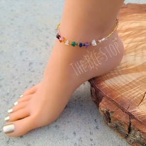 May include: A gold anklet with small, colorful gemstone beads. The anklet is on a model's ankle and the text "THE BEE STOP" is visible on the model's foot.