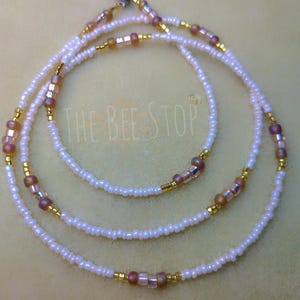 Puede incluir: Un collar de cuentas de cintura de tres hebras con cuentas blancas y doradas y detalles en rosa y dorado. Las cuentas están enhebradas en un cordón elástico transparente.
