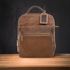 May include: A brown leather backpack with multiple zipped compartments and a top handle. The bag features a luggage tag and is set against a dark background on a wooden surface. The bag's dimensions are approximately 30 cm wide, 41 cm tall, and 15 cm deep.